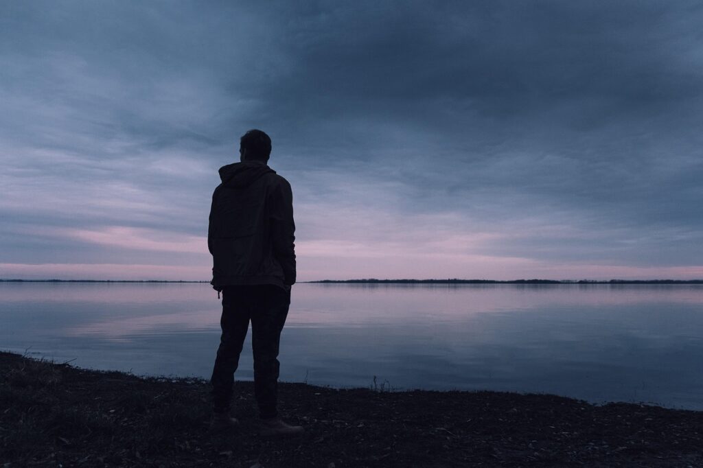 夕暮れの湖を前に一人で立ち尽くす男性のシルエット
