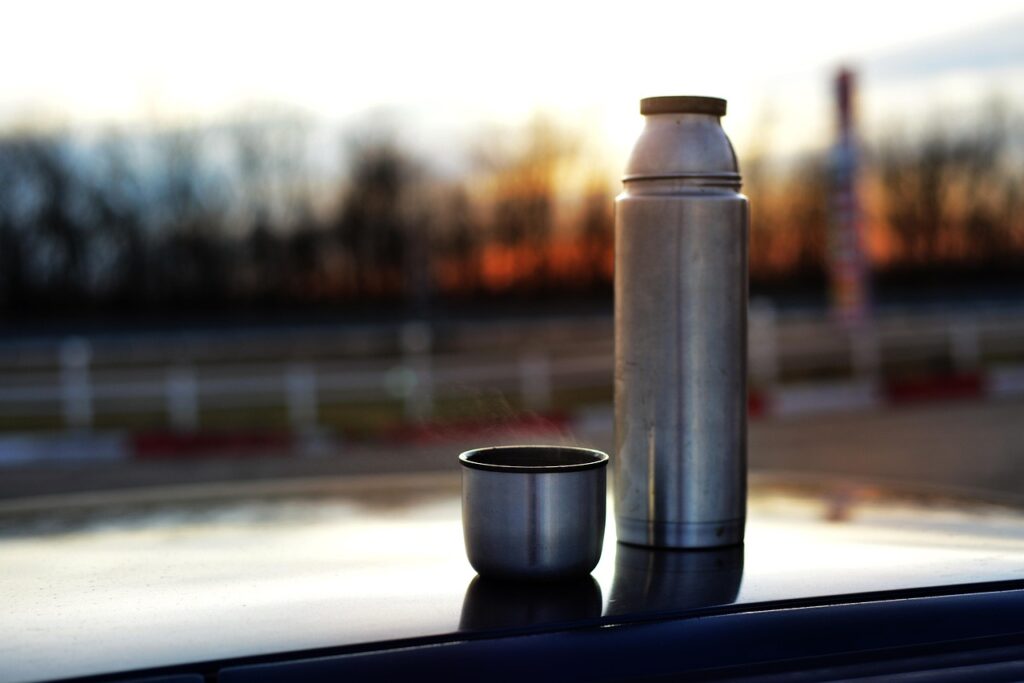 夕暮れの景色を背景に置かれたステンレスボトルと温かい飲み物の入ったカップ